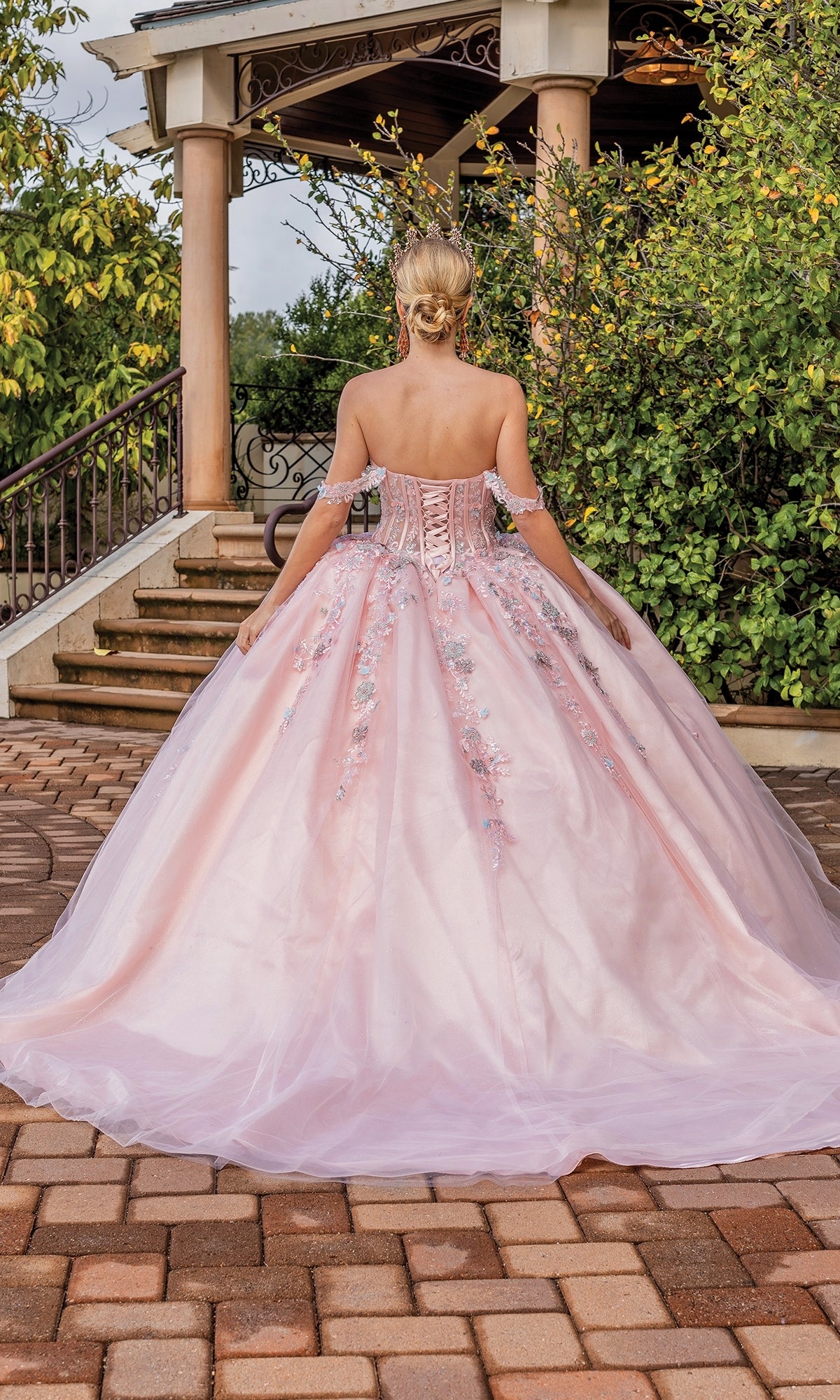 A woman stands with her back to the camera in front of a gazebo, wearing the blush pink Quinceanera Dress 1836 By Dancing Queen, featuring floral embellishments and an off-the-shoulder design.